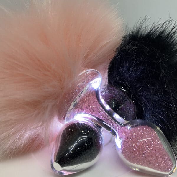 Bild von zwei übereinander angeordneten Plugs, der eine pink, der andere schwarz, gefüllt mit leuchtenden Perlen in ihren jeweiligen Farbtönen, jeder mit einem hängenden Hasenschwanz verziert.