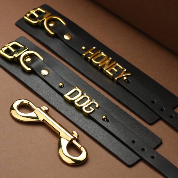 Ein Paar Handschellen aus weichem, schwarzem, italienischem Leder mit den Worten DOG und HONEY in 24 Karat vergoldeten Buchstaben mit eingearbeiteten Steinen auf jedem Buchstaben. Das Ganze liegt flach auf einem braunen Hintergrund mit einem Licht aus der oberen linken Ecke des Bildes;das Bild ist eine Idee für die Handschellen der Zusammenarbeit UPKO X Brigade Mondaine