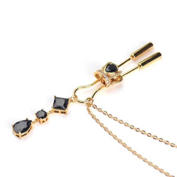 Crown breast clips from Upko. The product is gold-colored. Part of the product is a gold breast clamp with a rhinestone crown at the bottom and a black stone in the center. At the end is a gold chain with three black stones and the beginning of a double gold chain.