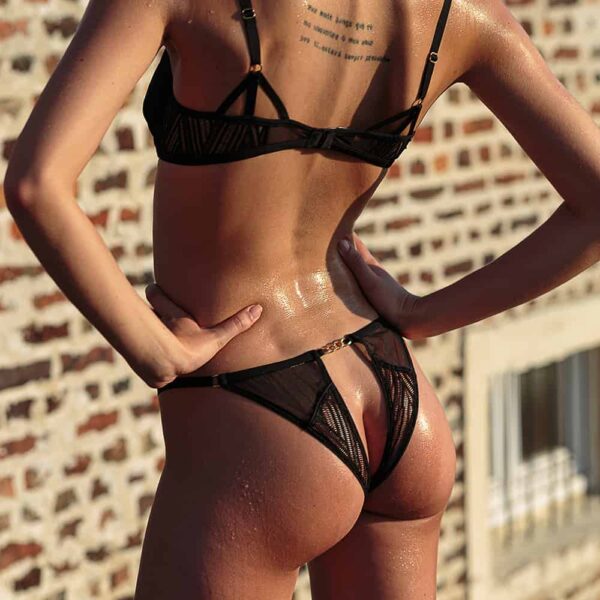 Черные открытые трусики и бюстгальтер от Onde Sensuelle из Atelier Amour в наличии в Brigade Mondaine. Трусики прозрачные с этническими узорами, за исключением концов. По поясу трусиков сзади проходит цепочка черного цвета. Бюстгальтер имеет такой же рисунок и цепочки на бретелях.