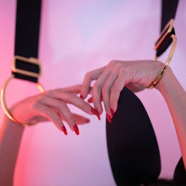 Varnished women's hands, held above her head by wide black straps with gold fastenings at the ends. The wrists are fitted with gold rings.