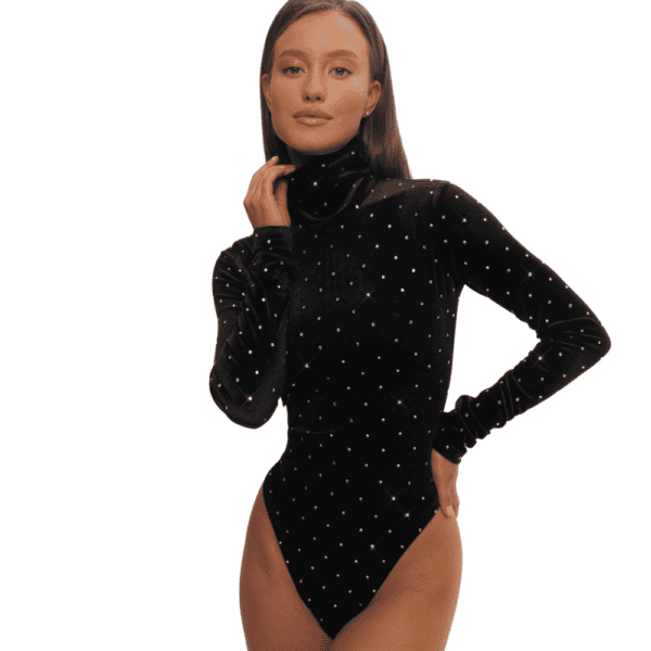 Una mujer posa elegantemente frente a una pared blanca, vestida con un body de terciopelo negro y cuello alto. Esta sofisticada prenda se ve realzada por la meticulosa incrustación de cristales de Swarovski, que añaden un toque de brillo y resplandor a su atuendo. La combinación de terciopelo oscuro y cristales brillantes crea un cautivador contraste visual, subrayando la sofisticación y la atención al detalle en sus elecciones de vestuario. Esta combinación única de texturas y elementos decorativos confiere al conjunto un aire chic y moderno, captando la atención y reflejando el refinado gusto de una mujer por la moda.