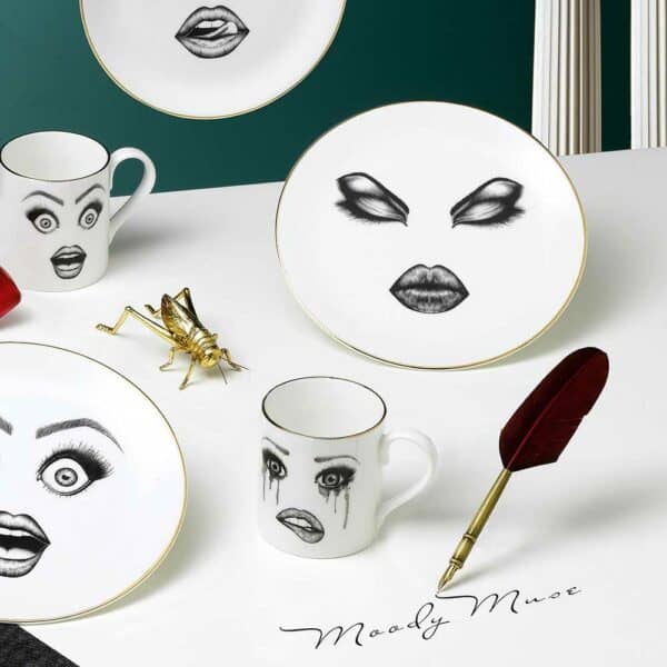 Moody Muse - Lauren Dickinson Clarke - Brigade Mondaine Eine Geschirrkollektion mit personalisierten Gesichtern und fesselnden Gesichtsausdrücken erwacht auf einem weißen Tisch zum Leben und kontrastiert wunderschön mit dem blauen Wandhintergrund.