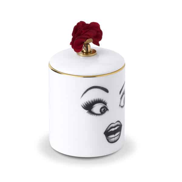 LDC - Prankster Duftkerze Kerze Muse Parfumée Ein überraschtes Gesicht, zart mit Filz auf weißes Porzellan mit goldenen und roten Details gezeichnet, auf einem weißen Tisch vor einer rosafarbenen Wand, auf der eine Ballerina mit einer Schleife thront.