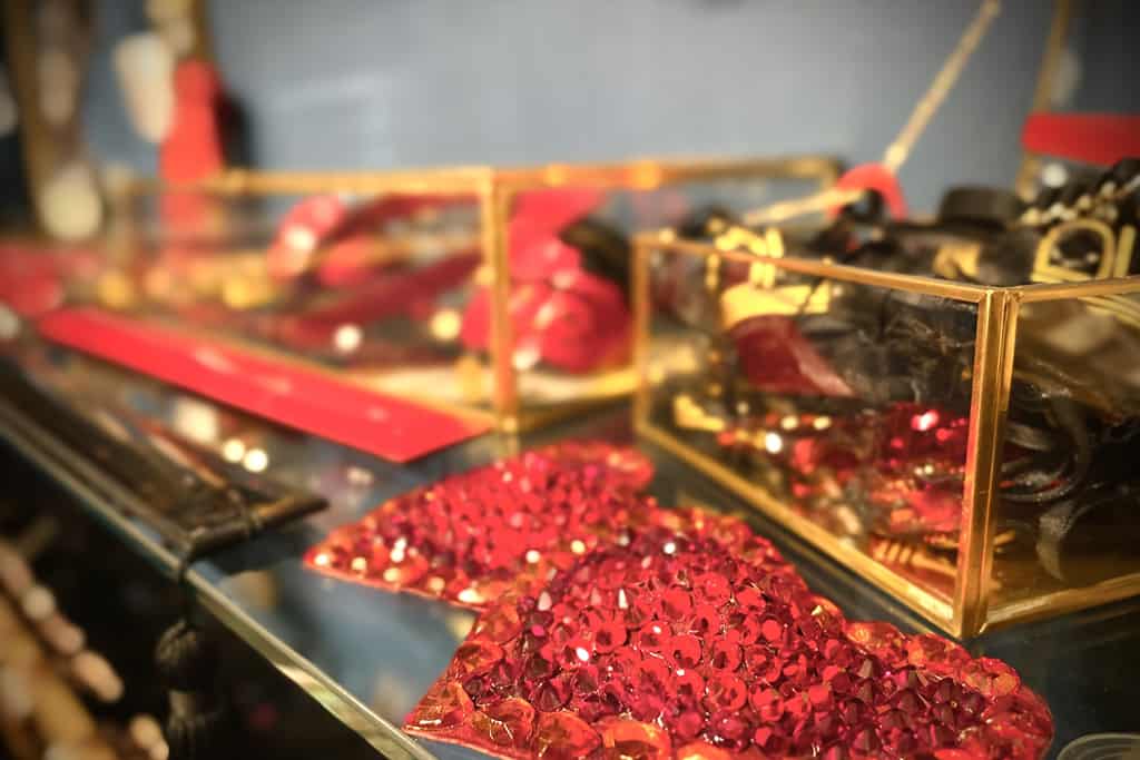 A close-up photograph shows two transparent boxes adorned with gold details. In the foreground are two red hearts encrusted with rhinestones. Inside the boxes are red objects and black pockets.
