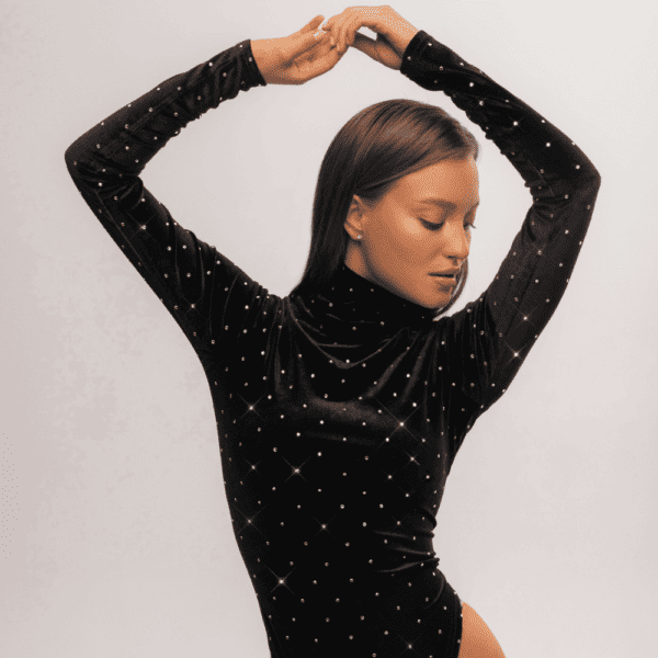 Una mujer posa elegantemente frente a una pared blanca, vestida con un body de terciopelo negro y cuello alto. Esta sofisticada prenda se ve realzada por la meticulosa incrustación de cristales de Swarovski, que añaden un toque de brillo y resplandor a su atuendo. La combinación de terciopelo oscuro y cristales brillantes crea un cautivador contraste visual, subrayando la sofisticación y la atención al detalle en sus elecciones de vestuario. Esta combinación única de texturas y elementos decorativos confiere al conjunto un aire chic y moderno, captando la atención y reflejando el refinado gusto de una mujer por la moda.