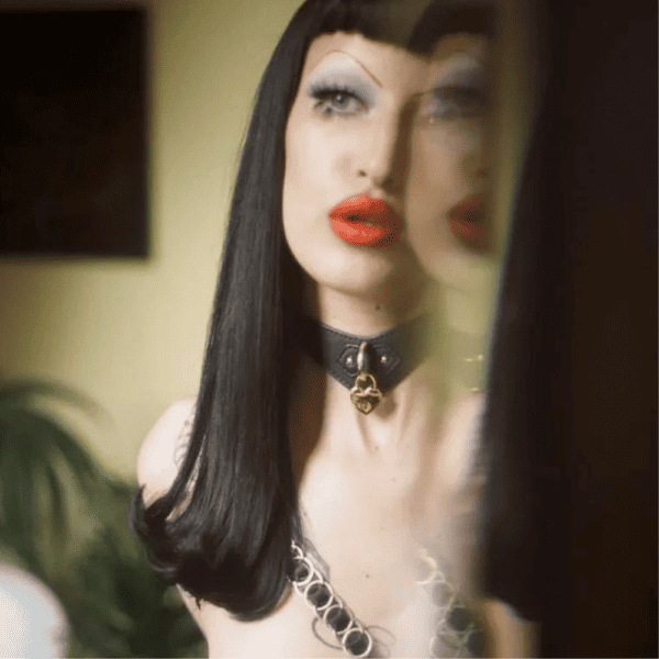 Fotografía tomada en el reflejo de un espejo, una mujer morena con flequillo corto, faja fina y maquillada lleva un collar de cuero negro con un candado de corazón.
