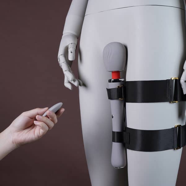 Bild zeigt ein Bein, an dem ein Set mit einem schwarzen Geschirr sowie einem weißen Vibrator befestigt ist. Eine Hand hält die Fernbedienung.