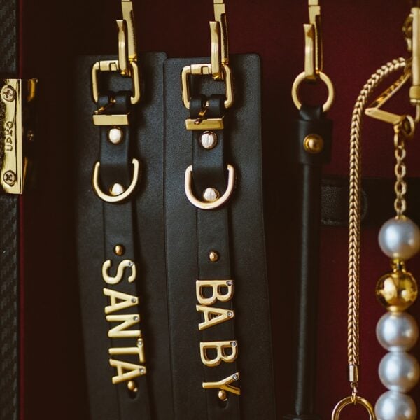 UPKO accessories in black leather with adjustable elastics, mother-of-pearl beads and gold metal finishes, including collars, handcuffs and custom pieces, presented on a burgundy background, available at Brigade Mondaine Paris.
