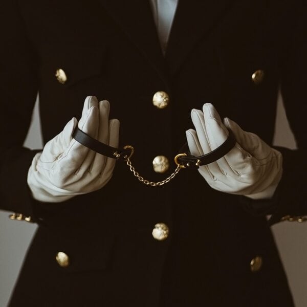 Menottes UPKO en cuir noir avec chaîne dorée, portées sur gants blancs, sélection Brigade Mondaine Paris