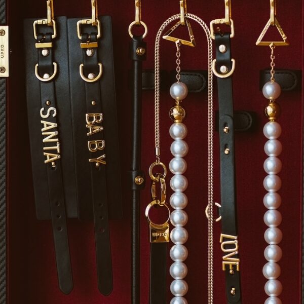UPKO accessories in black leather with adjustable elastics, mother-of-pearl beads and gold metal finishes, including collars, handcuffs and custom pieces, presented on a burgundy background, available at Brigade Mondaine Paris.