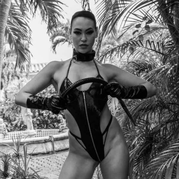 Foto de una mujer con un body de cuero negro «Serpens», un collar bondage de cuero de vaca negro con anilla metálica y una cola de impacto negra de cuero