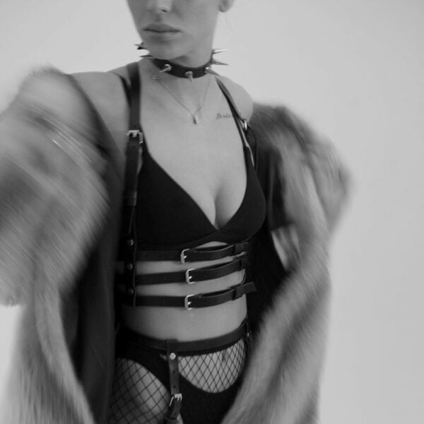 Woman in black and white wearing a handmade natural leather harness, with three straps under the bust and two straps rising to the neck, creating a bra effect. She's also wearing a black bra, and the leather is highlighted by the light, accentuating the texture and sophisticated design.
