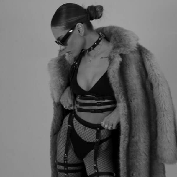 Woman in black and white wearing a handmade natural leather harness, with three straps under the bust and two straps rising to the neck, creating a bra effect. She's also wearing a black bra, and the leather is highlighted by the light, accentuating the texture and sophisticated design.