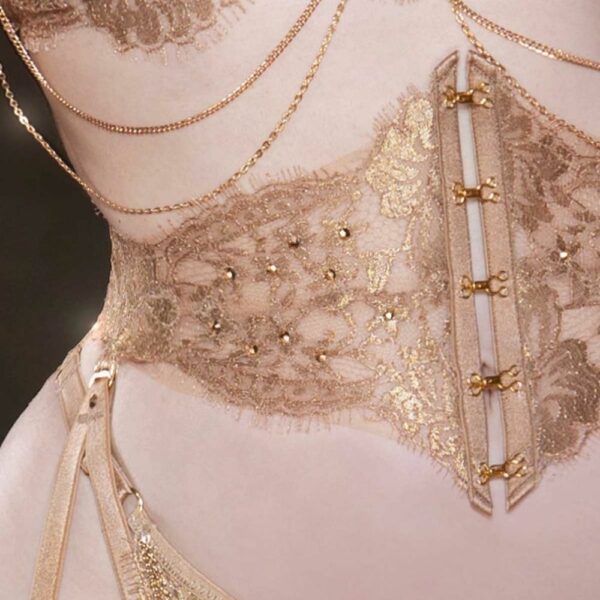 HERVÉ by Céline Marie AIMÉE - Korsett Strapsgürtel Das Korsett „Taille AIMÉE“ ist ein raffiniertes, handgefertigtes Meisterwerk aus französischer Leavers-Spitze in Roségold und ultratransparentem Tüll. Es ist mit goldenen Ringen und einer Schnürung am Rücken versehen, die eine individuelle Passform ermöglicht. Die Vorderseite zieren präzise aufgenähte goldene Haken und Ösen, und der mit flexiblen Stäbchen strukturierte Unterbau formt eine perfekte Silhouette. Die abnehmbaren doppelten Träger sorgen für zusätzlichen Komfort und Flexibilität. Es kann als Taillenband oder Strumpfhalter getragen werden und lässt sich mit dem Bralette oder dem BH aus derselben Kollektion kombinieren.