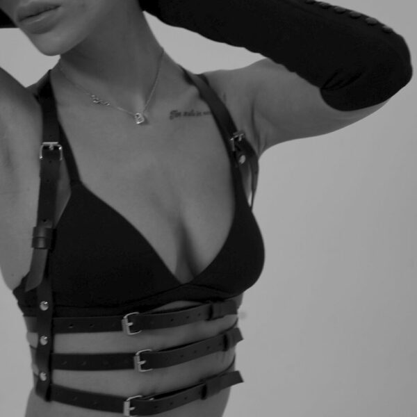 Woman in black and white wearing a handmade natural leather harness, with three straps under the bust and two straps rising to the neck, creating a bra effect. She's also wearing a black bra, and the leather is highlighted by the light, accentuating the texture and sophisticated design.