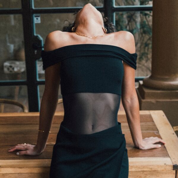 Undress Code – Body Libra | Brigade Mondaine Paris Body noir Undress Code Libra à épaules dégagées, porté par un modèle tenant une coupe de champagne, sélection Brigade Mondaine Paris.