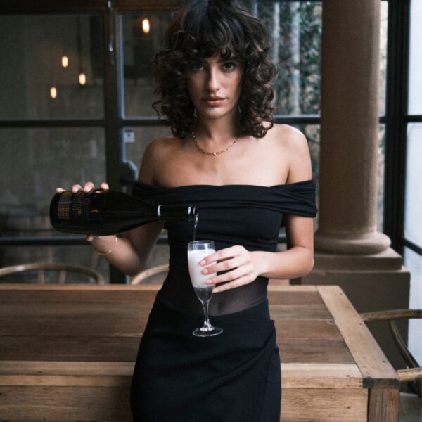 Undress Code – Body Libra | Brigade Mondaine Paris Body noir Undress Code Libra à épaules dégagées, porté par un modèle tenant une coupe de champagne, sélection Brigade Mondaine Paris.