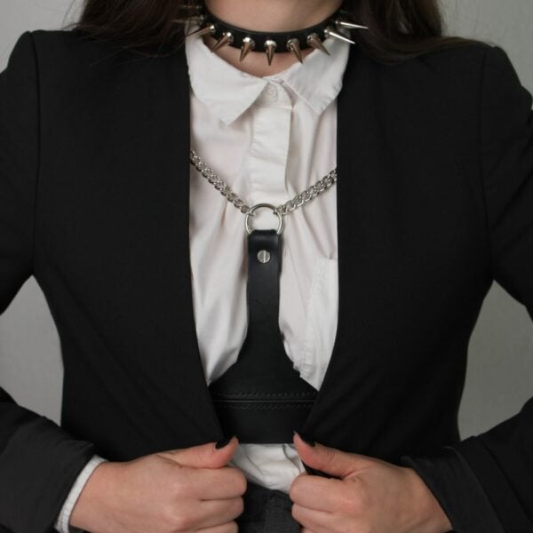NIAR House - Choker 86137 Frau trägt den Choker 86137 mit weißem Hemd, schwarzem Blazer und schwarzem Ledergeschirr, das mit silbernen Ketten verziert ist. Der Choker ist mit Metallspikes verziert und wird hinten mit einer verstellbaren Schnalle geschlossen.