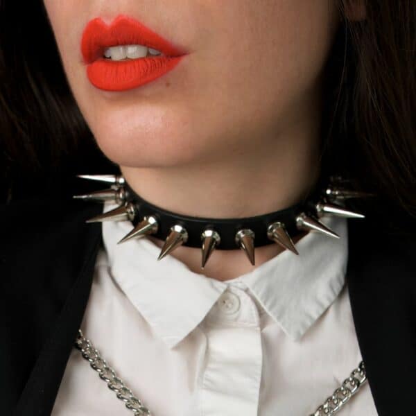 NIAR House - Choker 86137 Nahaufnahme des Chokers 86137, getragen von einer Frau mit rotem Lippenstift, weißem Hemd und schwarzem Blazer, der mit zwei silbernen Ketten verziert ist. Der Choker ist mit 26 mm langen und 1,5 cm breiten Metallspikes verziert und hat eine verstellbare Passform auf der Rückseite.