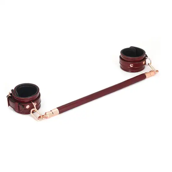 Spreizstange aus weinrotem Leder mit einem Kern aus Eukalyptusholz. O-Ringe aus roségoldfarbenem Metall zur Befestigung von Knöchelschienen. Elegant und funktional für raffinierte BDSM-Spiele.