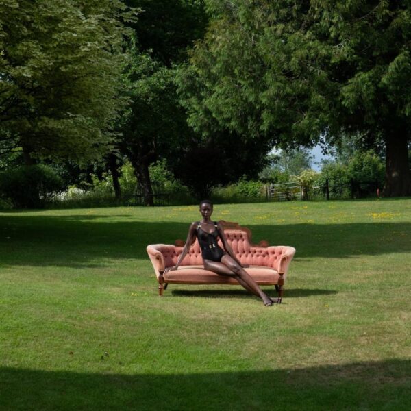 BORDELLE – Body Noir Sur Canapé Ancien En Plein Jardin Mannequin assis sur un canapé ancien rose poudré posé sur une pelouse, portant un body noir BORDELLE, dans un vaste jardin arboré.