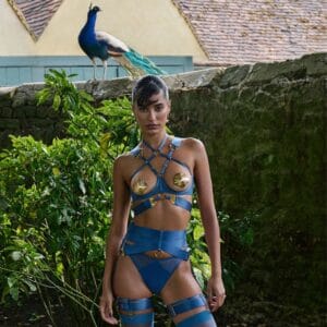 Model in einem blauen BORDELLE-Ensemble aus elastischen Bändern, goldenen Muschelverzierungen und hochgeschnittenen Strümpfen, fotografiert in einem Garten mit einem Pfau, der auf einer Steinmauer im Hintergrund sitzt.