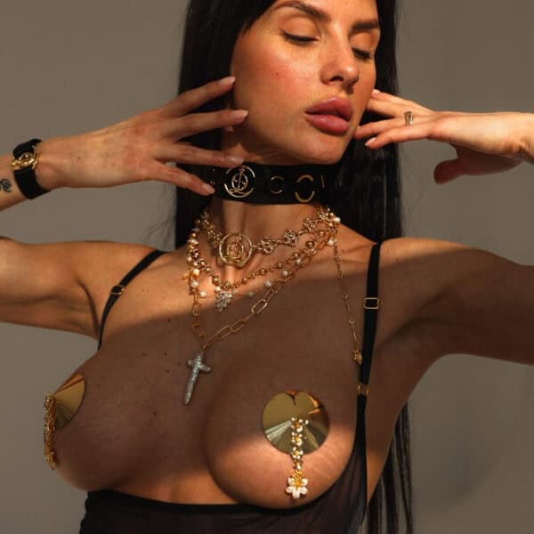 Goldener Schmuck und Nipple Covers QRUCIFIX - BRIGADE MONDAINE - Luxus-Dessous Paris Frau, die mehrere goldene QRUCIFIX-Ketten und scheibenförmige Metall-Nippelcovers mit Anhängern trägt. Sie posiert mit erhobenen Armen und geschlossenen Augen. Schwarzer Choker mit Logo und sichtbaren schwarzen Dessous-Trägern.