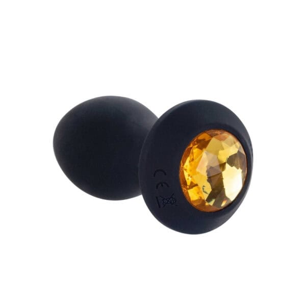 Schwarzer Analplug aus Silikon mit einer Basis, die mit einem facettierten bernsteinfarbenen Schmuckstück verziert ist, in der Hand gehalten, angeboten von Brigade Mondaine Paris.