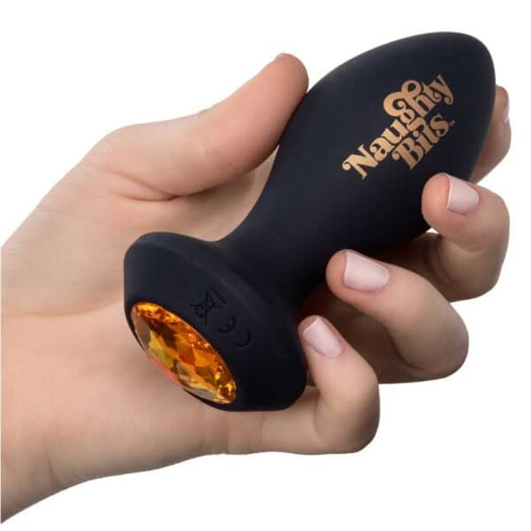 Schwarzer Analplug aus Silikon mit einer Basis, die mit einem facettierten bernsteinfarbenen Schmuckstück verziert ist, in der Hand gehalten, angeboten von Brigade Mondaine Paris.