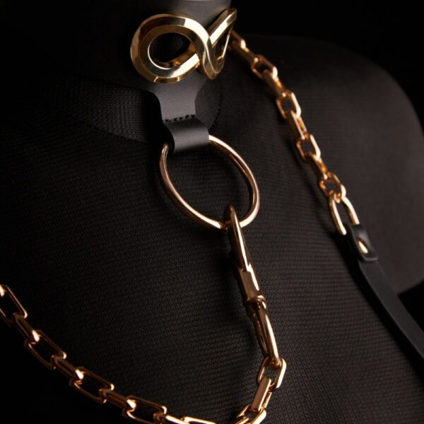 Halsband Leine SEVERA IINFYNII - Brigade Mondaine Paris Die Leine SEVERA von IINFYNII kombiniert schwarzes Leder und eine goldene Metallkette in einer selbstbewussten und strukturierenden Konstruktion. Sie ist als starkes Körperteil gedacht und besteht aus einem Lederhalsband, das mit einem Signatur-Metallsymbol verziert ist und durch eine Gelenkkette verlängert wird, die mit einem zentralen Ring verbunden ist. Es ist anpassbar und kann allein getragen oder in eine komplette Silhouette integriert werden, zwischen Schmuckstück mit Zwangscharakter und skulpturalem Accessoire. Ein grafisches, direktes Stück, das für diejenigen entworfen wurde, die ihren Körper als ein Feld des Ausdrucks betrachten.
