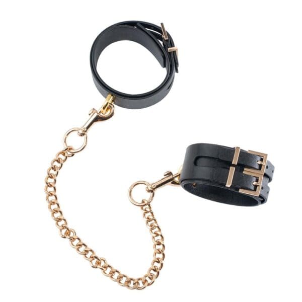 Esposas tobilleras premium en cuero negro | Brigade Mondaine Par de tobilleras de piel negra unidas por una cadena dorada, con hebillas metálicas ajustables y meticulosamente acabadas, presentadas sobre un fondo blanco de estética refinada.