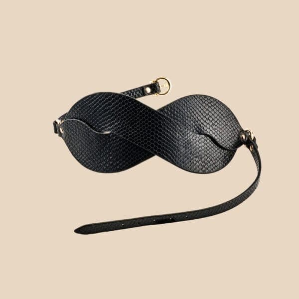 Masque BDSM en cuir noir texturé à la forme enveloppante, avec attaches réglables et bouclerie dorée, présenté sur fond noir dans une mise en scène élégante et minimaliste.