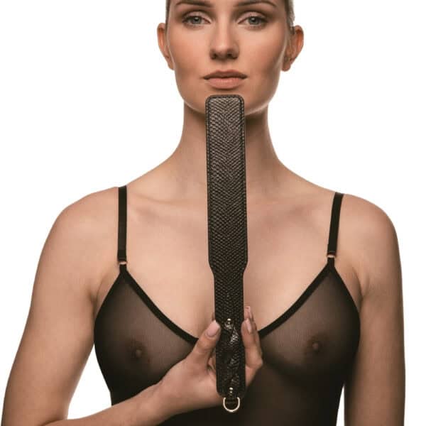 Model trägt einen transparenten schwarzen Body und hält ein BDSM-Paddle aus schwarzem Leder mit strukturiertem Finish. Fotografiert vor einem weißen Hintergrund in einer schlichten und eleganten Inszenierung.