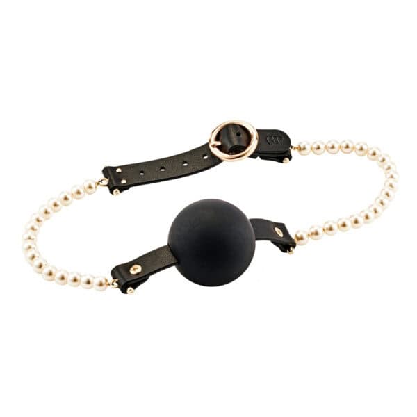 Mordaza de bola de silicona negra con correas de cuero y perlas blancas, presentada plana sobre fondo claro, ofrecida por Brigade Mondaine Paris.