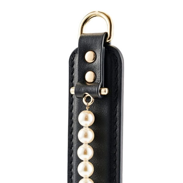 Paddle BDSM en cuir noir avec finition lisse et rangée de perles décoratives, tenu par un modèle en studio, accessoire sélectionné par Brigade Mondaine Paris.