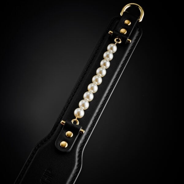 Paddle BDSM en cuir noir avec finition lisse et rangée de perles décoratives, tenu par un modèle en studio, accessoire sélectionné par Brigade Mondaine Paris.