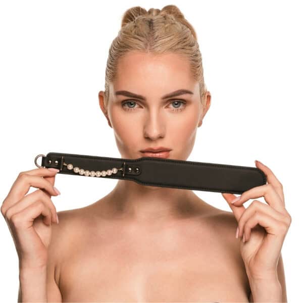Paddle BDSM en cuir noir avec finition lisse et rangée de perles décoratives, tenu par un modèle en studio, accessoire sélectionné par Brigade Mondaine Paris.