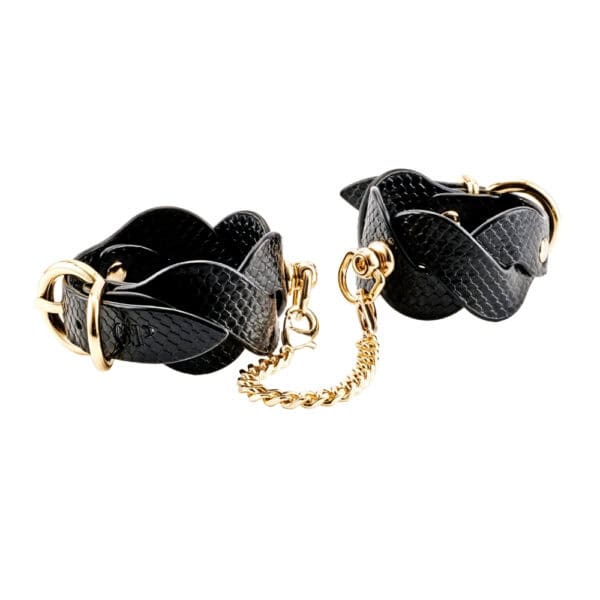 Chevilles d’un modèle portant des menottes BDSM noires texturées reliées par une chaîne dorée, accessoire de contrainte sélectionné par Brigade Mondaine Paris.