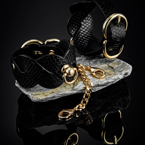 Modèle portant des menottes BDSM noires pour poignets avec chaîne dorée, accessoire de contrainte proposé par Brigade Mondaine Paris.