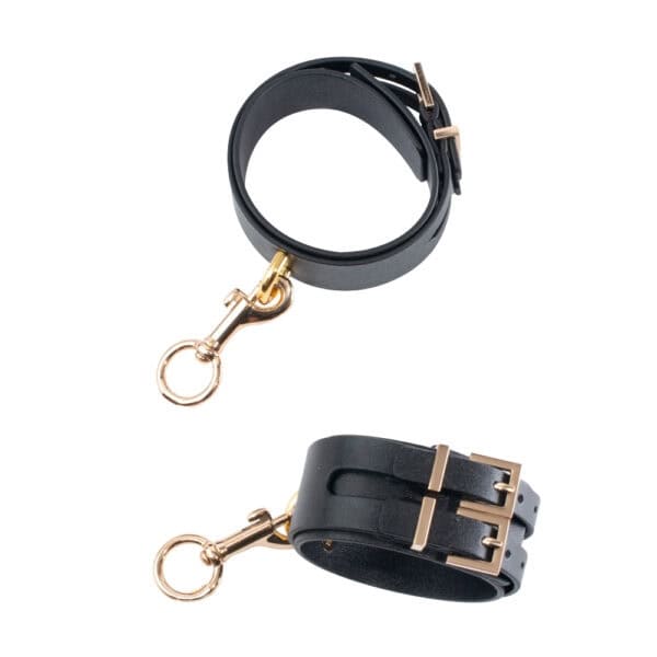 BDSM Fußfesseln aus schwarzem Leder mit goldener Kette | Brigade Mondaine BDSM-Fußfesseln aus schwarzem Leder mit Metallschnalle und goldener Kette, die an den Fußgelenken zu schwarzen Pumps mit Absätzen getragen werden, fotografiert vor einem grauen Hintergrund.