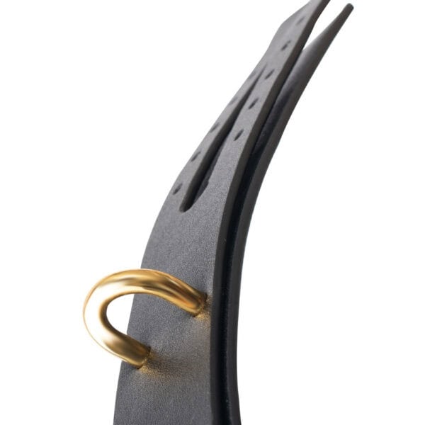 Esposas tobillo BDSM cuero negro y cadena de oro | Brigade Mondaine Esposas tobilleras BDSM de cuero negro con hebilla metálica y cadena dorada, llevadas en los tobillos con zapatos de tacón negros, fotografiadas sobre fondo gris.