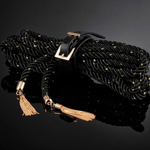 BDSM Seil schwarz Luxus goldene Details | Brigade Mondaine Schwarzes, gedrehtes BDSM-Seil mit goldenen Einlagen, das mit einem Lederriemen und einer Metallschnalle geschlossen wird, mit goldenen Verzierungen und Pompons versehen ist und auf schwarzem Hintergrund präsentiert wird.