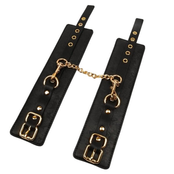 BDSM-Set aus schwarzem Leder, bestehend aus einem Paar Handschellen, die mit einer goldenen Kette verbunden sind, einer Verdunkelungsmaske und einer passenden Reitgerte, präsentiert auf weißem Hintergrund in einer eleganten Inszenierung.
