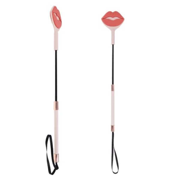 Látigo BDSM con diseño decorativo de labios, mango delgado y correa para la muñeca, presentado por Brigade Mondaine Paris sobre fondo claro.