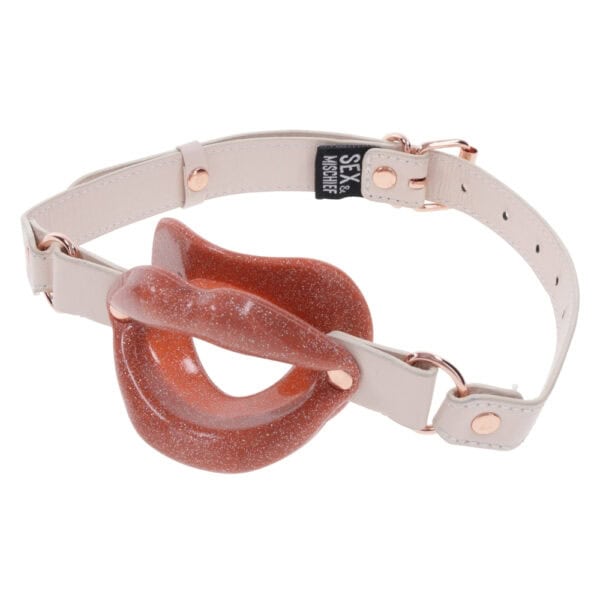 Open gag in lip design with clear strap and metal finishes, presented by Brigade Mondaine Paris on clear background.