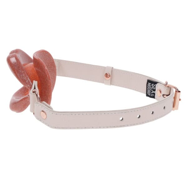 Open gag in lip design with clear strap and metal finishes, presented by Brigade Mondaine Paris on clear background.