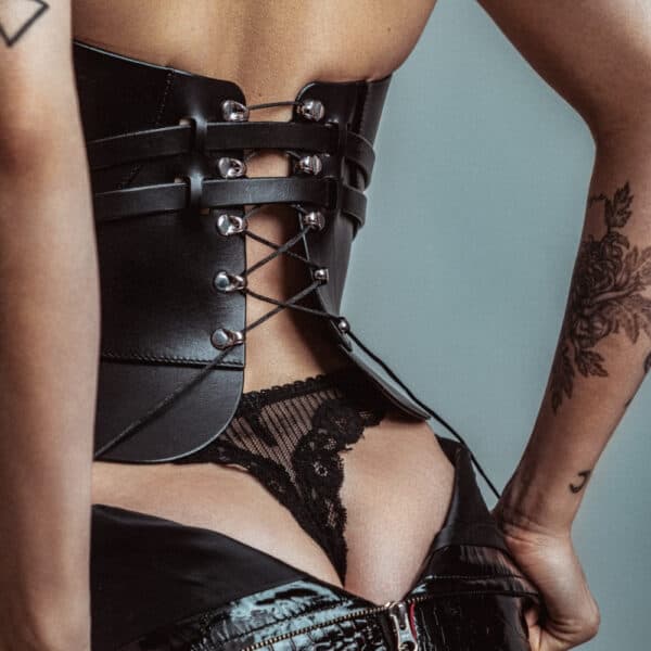 Corsé de cuero Daelos RE:STRICT Brigade Mondaine Paris Modelo con corsé Daelos de piel negra RE:STRICT en Brigade Mondaine Paris, escote pronunciado estructurado, tirantes ajustables, con accesorios a juego sobre fondo rojo.