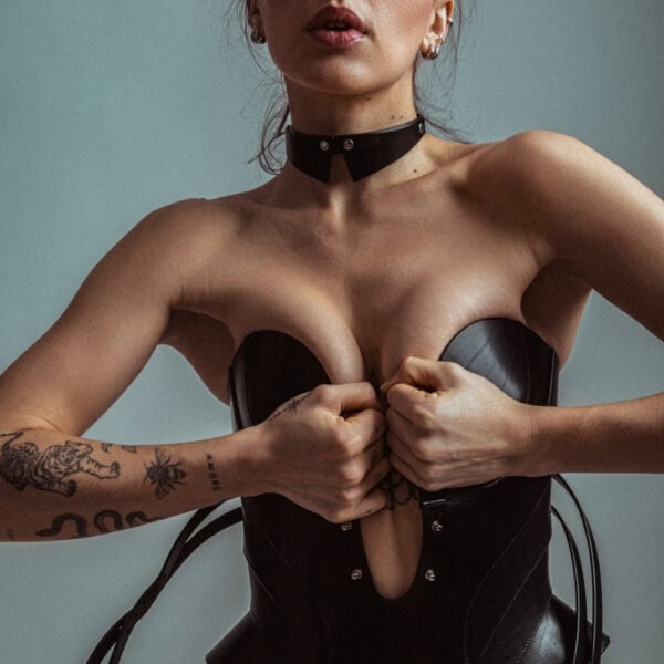 Corsé de cuero Daelos RE:STRICT Brigade Mondaine Paris Modelo con corsé Daelos de piel negra RE:STRICT en Brigade Mondaine Paris, escote pronunciado estructurado, tirantes ajustables, con accesorios a juego sobre fondo rojo.