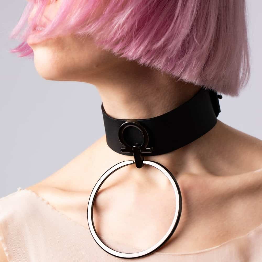 Modell trägt den Divine Perlensau Choker aus schwarzem Leder mit einem breiten, hängenden Metallring, angeboten von Brigade Mondaine Paris.